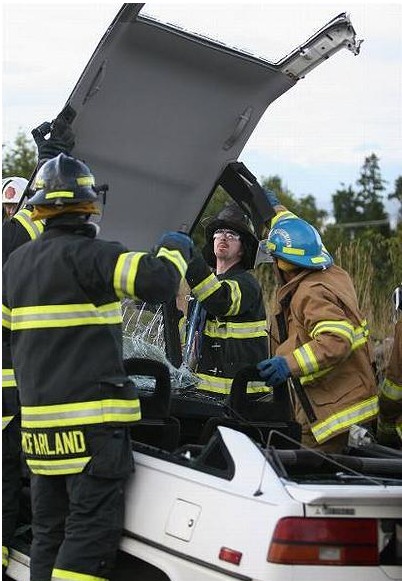 Firefigters in bunker gear and helmets remove the top of a white car as practice for an extrication of an injured occupant.